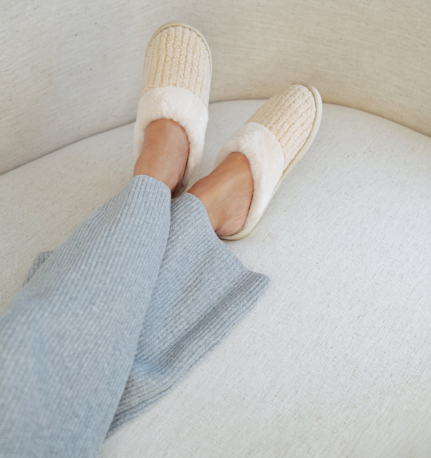 Person wearing white slippers on a light-colored couch. $10 slipper with $99 purchase